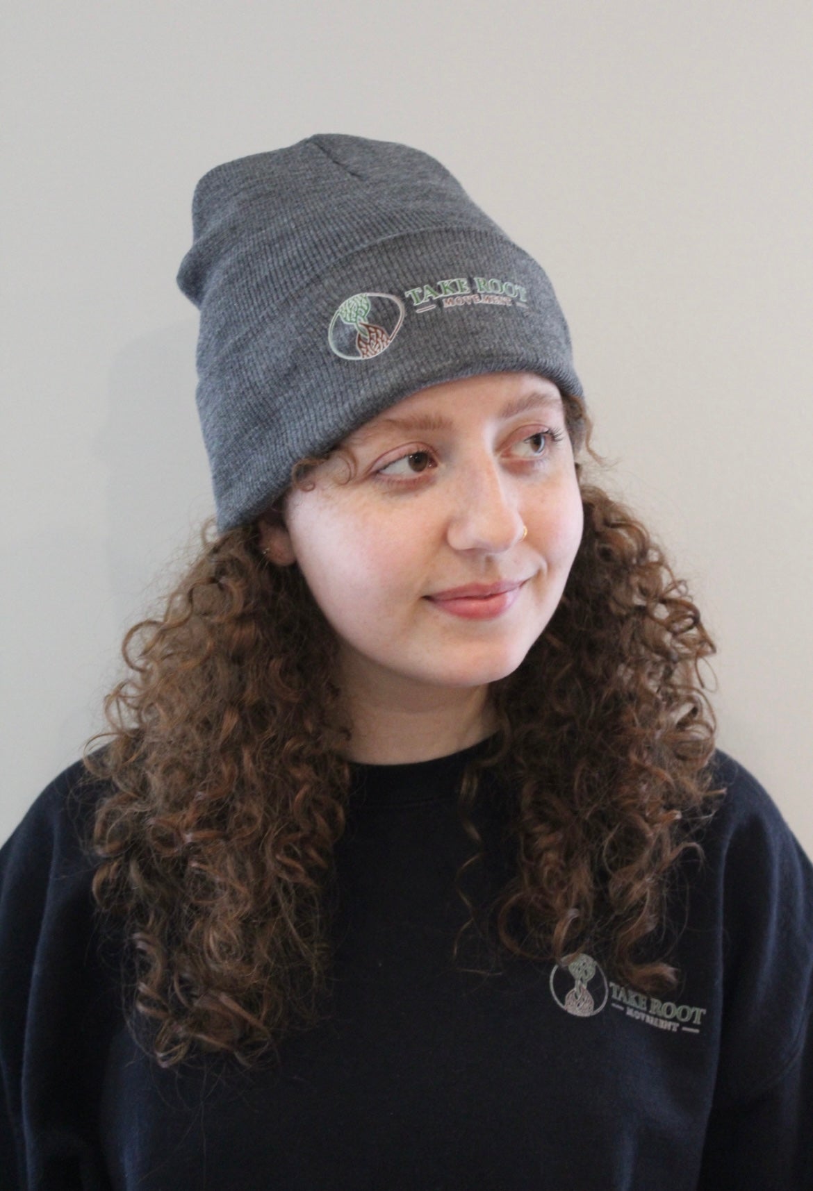 Female wearing dark Grey beanie with hour glass logo with Take Root Movement next to it