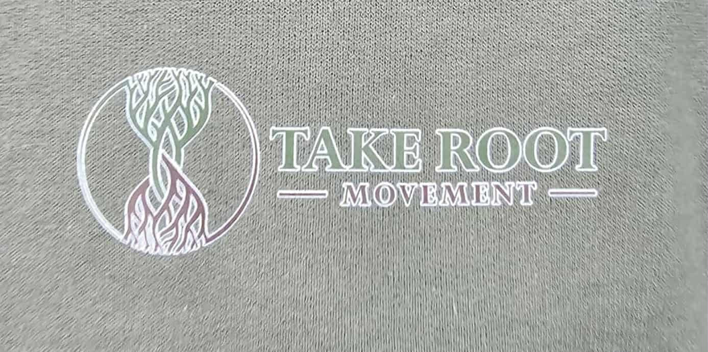 Close-up of Take Root Movement logo with intertwined tree and roots symbol on olive green fabric.