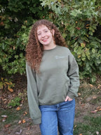 Person wearing olive green Take Root Movement sweatshirt with small tree-and-roots logo on the front left chest.