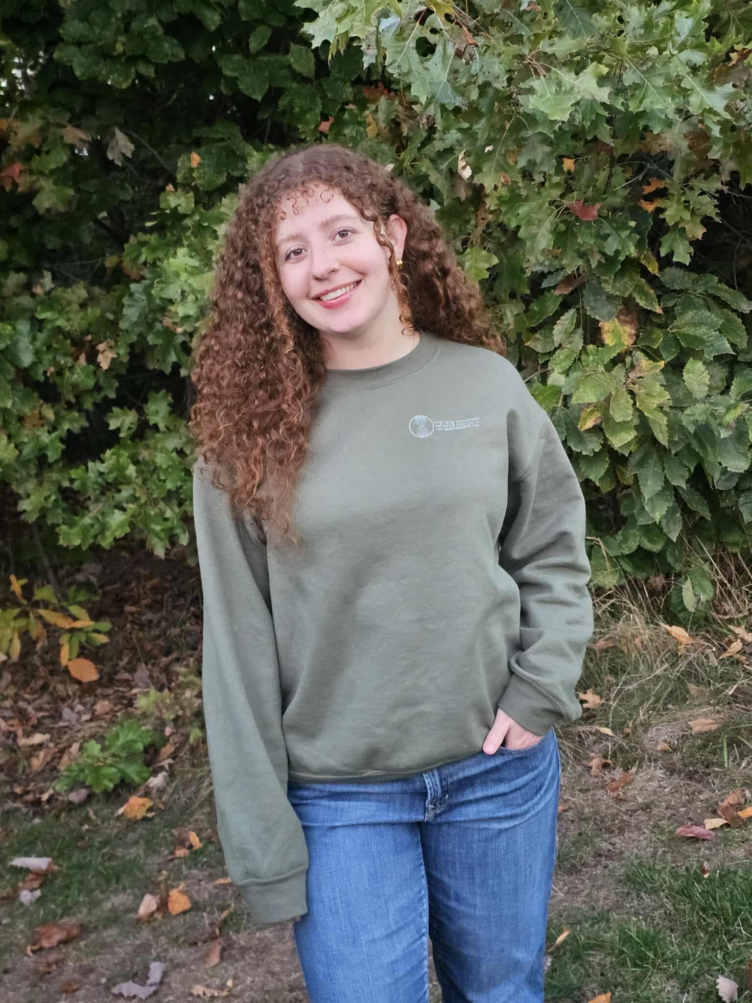Person wearing olive green Take Root Movement sweatshirt with small tree-and-roots logo on the front left chest.