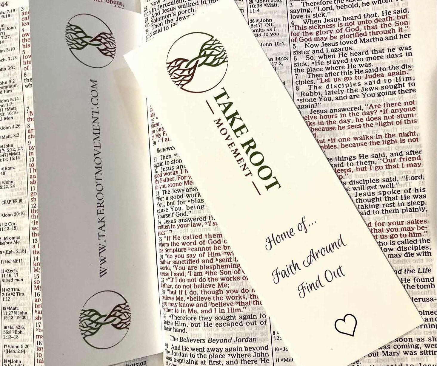 Two Take Root Movement Christian bookmarks placed inside an open Bible, featuring the intertwined tree and roots logo, ‘Faith Around Find Out’ message, and website on white cardstock.