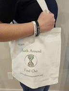 Person carrying a white canvas Take Root Movement Christian tote bag with the ‘Faith Around Find Out’ tree and roots logo, wearing matching Faith Around silicone bracelets.