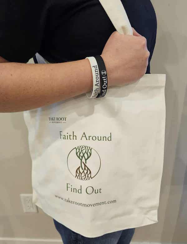 Person carrying a white canvas Take Root Movement Christian tote bag with the ‘Faith Around Find Out’ tree and roots logo, wearing matching Faith Around silicone bracelets.