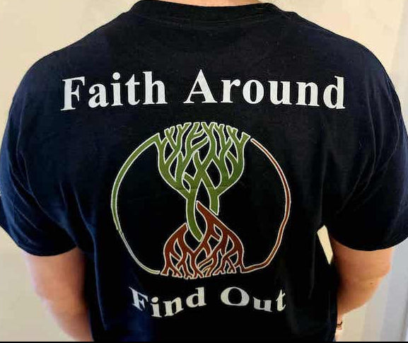 Person wearing black Take Root Movement Christian T-shirt with small tree and roots logo printed on the left chest, shown standing indoors.